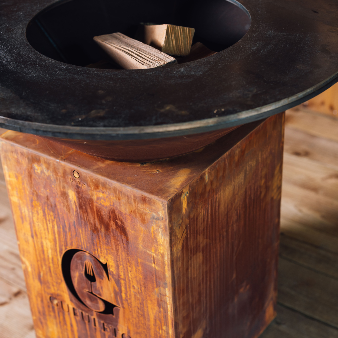 Gros plan sur le socle en acier Corten du brasero Classic 100 Gueuleton, avec le logo gravé et le bois empilé prêt à être allumé.