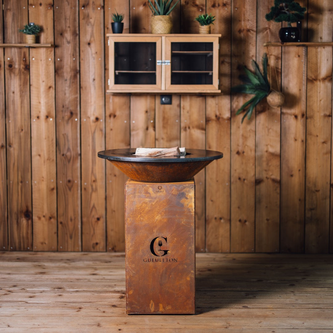 Brasero Gueuleton 75 en acier corten, posĂ© sur un sol en bois, avec une paroi en bois en arriĂšre-plan et du bois de chauffe prĂȘt Ă ĂȘtre allumĂ© sur la plaque de cuisson ronde.