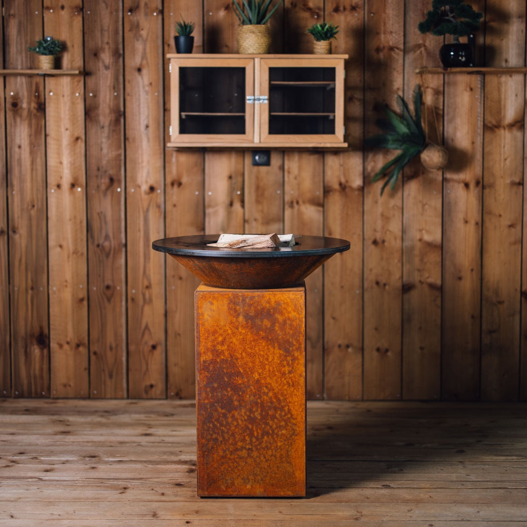 Vue arriÚre du brasero Gueuleton 75 en acier corten, posé sur un sol en bois avec un mur lambrissé en fond. La face arriÚre du socle ne comporte aucun logo, mettant en valeur la texture naturelle du métal patiné.
