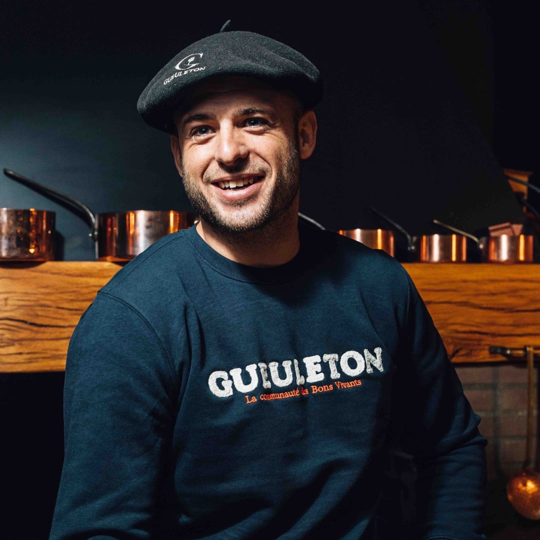 Homme souriant portant le béret Gueuleton noir brodé, dans une cuisine chaleureuse avec casseroles en cuivre en arrière-plan.