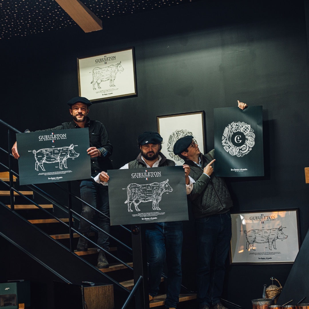 Maxime Huguet, Vincent Bernard et Arthur Edange avec des affiches Gueuleton montrant les découpes de viande de porc et de bœuf, sur fond de mur noir avec des cadres et illustrations de charcuterie. Affiches illustrant les morceaux de viande, une présentation authentique du savoir-faire de Gueuleton.