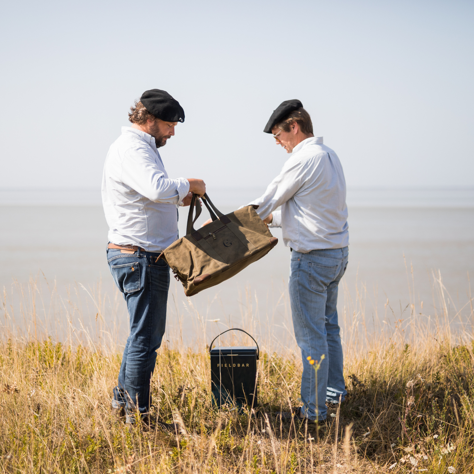 Deux hommes avec bérets échangeant un Sac de Voyage Vintage en toile cirée Héritage Gueuleton en plein air - Gueuleton