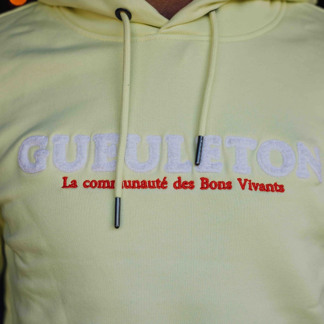 Zoom sur le logo brodé du sweat à capuche jaune Gueuleton avec l'inscription 'La communauté des Bons Vivants'