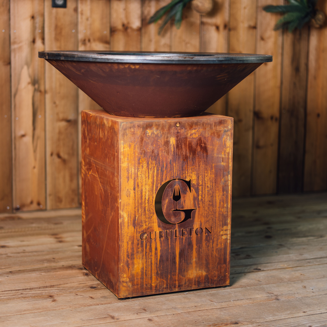Vue latérale du brasero Classic Corten 100 Gueuleton, avec cuve ouverte et piétement en acier rouillé, posé sur un parquet bois et fond mural en lames verticales.