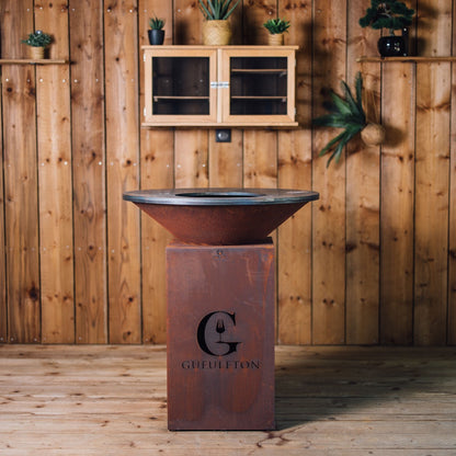 Vue frontale du brasero en acier Corten avec le logo Gueuleton centré, posé sur un parquet de bois avec un fond mural en planches verticales.