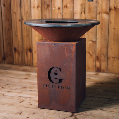 Brasero Classic Corten 85 en acier Corten, avec son logo Gueuleton découpé sur la face avant, posé sur un sol en bois devant un mur en lames de bois.
