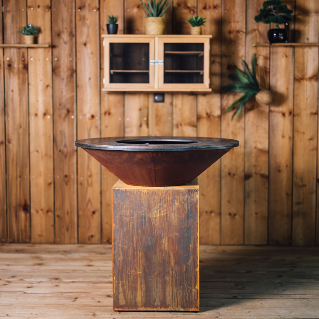 Vue arrière du brasero en acier Corten modèle Classic 100, avec son piétement patiné et son large foyer circulaire, dans un décor chaleureux en bois naturel
