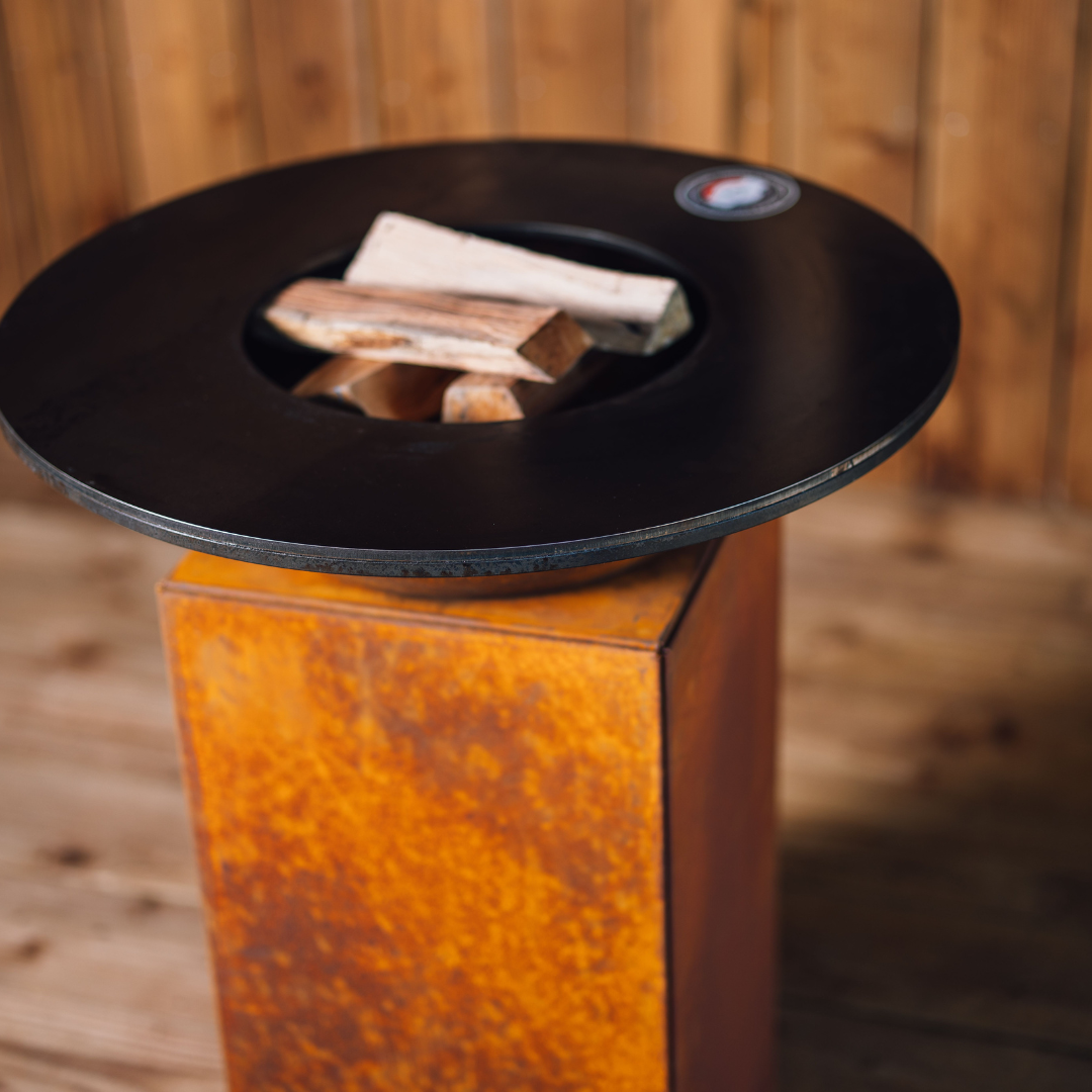Plan incliné du brasero Gueuleton 75 montrant la plaque de cuisson noire avec bois de chauffe au centre, sur socle en acier corten.
