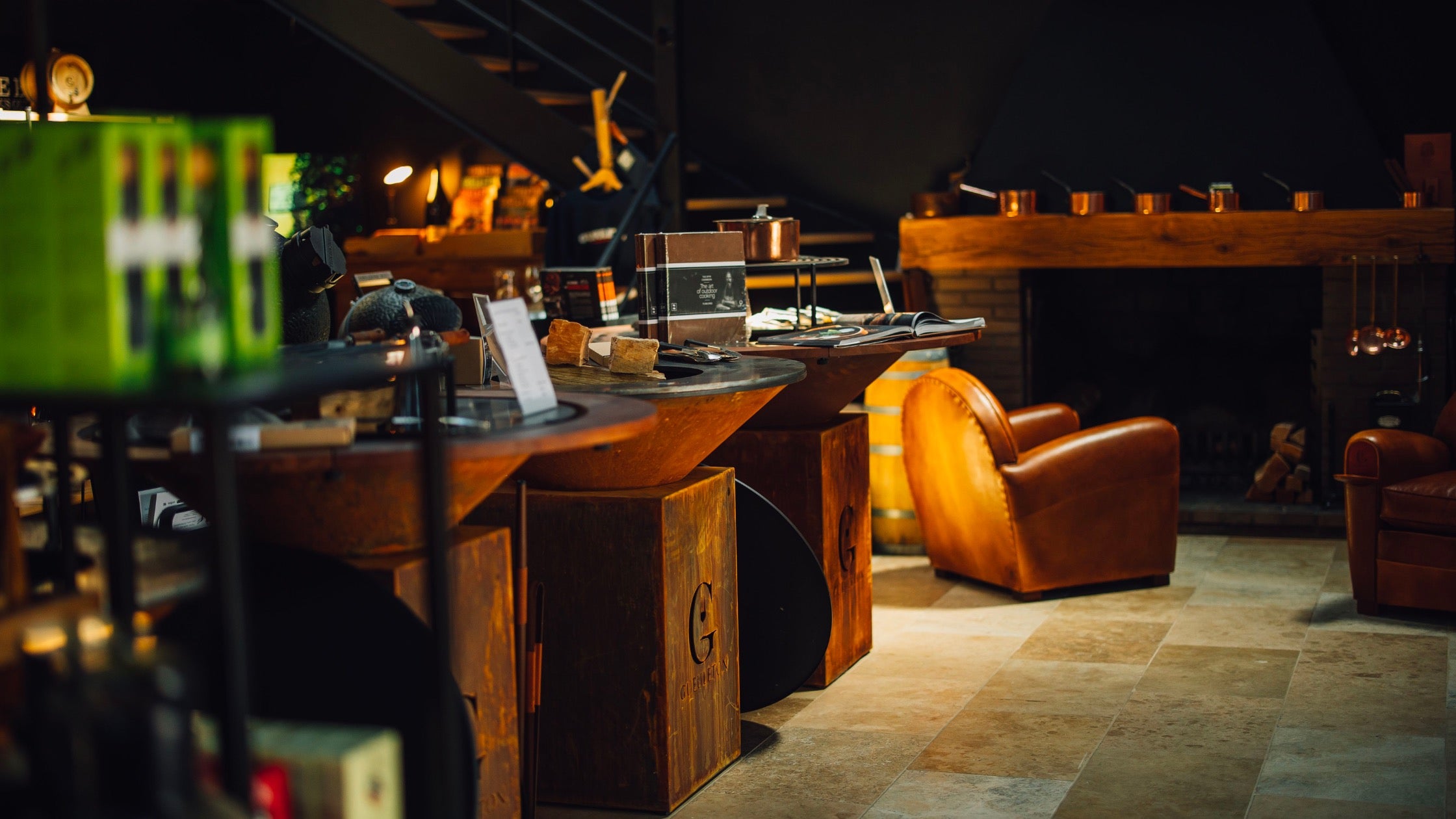 Vue du showroom Gueuleton avec deux braseros en acier Corten utilisés en présentoirs, fauteuils en cuir, cheminée rustique et ambiance chaleureuse.