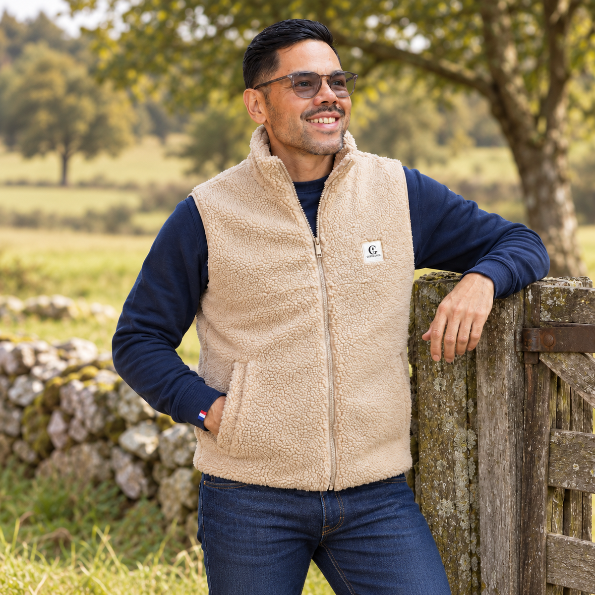 homme portant un gilet sherpa gueuleton sable a col montant sur un sweat blue marine, appuyé contre un portail.