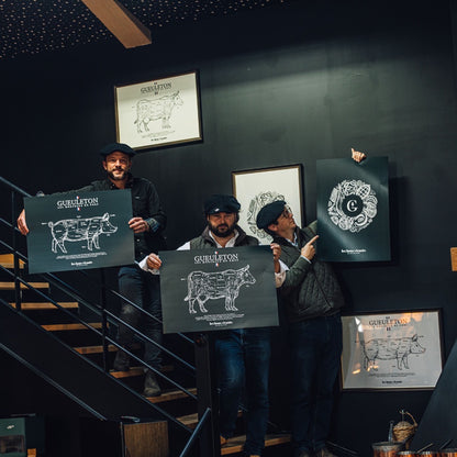 Maxime Huguet, Vincent Bernard et Arthur Edange avec des affiches Gueuleton montrant les découpes de viande de porc et de bœuf, sur fond de mur noir avec des cadres et illustrations de charcuterie. Affiches illustrant les morceaux de viande, une présentation authentique du savoir-faire de Gueuleton.