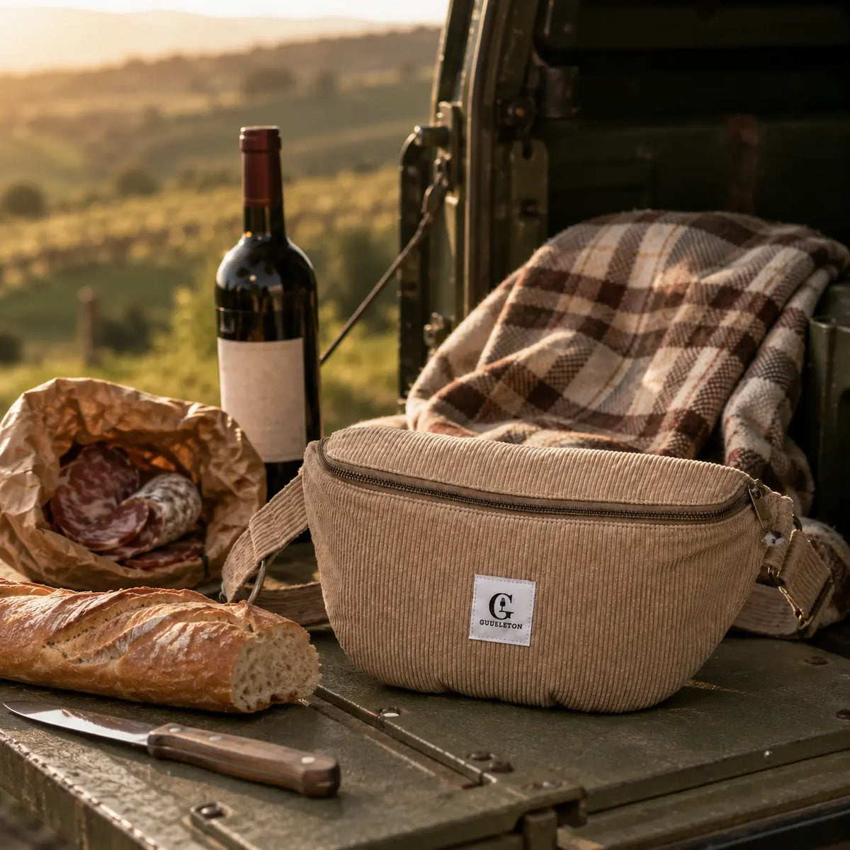 Sac banane Gueuleton beige sur le hayon d'un 4x4 avec baguette, saucisson et bouteille de vin